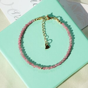 Op de afbeelding: Een delicate armband van roze edelstenen met een gouden hartbedel en een gouden sluiting. De armband wordt getoond op een lichtgroene achtergrond.