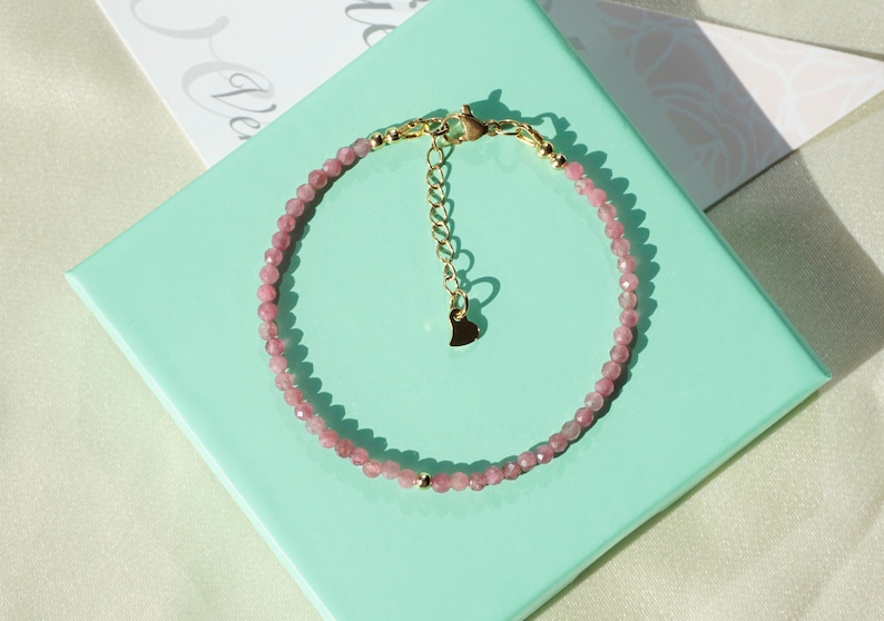 Op de afbeelding: Een delicate armband van roze edelstenen met een gouden hartbedel en een gouden sluiting. De armband wordt getoond op een lichtgroene achtergrond.