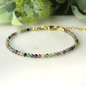 Könnte beinhalten: Ein zartes Armband mit kleinen, runden, mehrfarbigen Turmalinperlen in Grün-, Rosa- und Klartönen. Das Armband verfügt über einen goldfarbenen Karabinerverschluss aus Metall und eine verstellbare Verlängerungskette mit einem kleinen runden Anhänger.