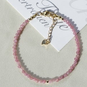 Op de afbeelding: Een delicate armband van roze edelstenen met een gouden sluiting en verlengketting. De armband is gemaakt van kleine, gefacetteerde kralen en heeft een hartvormige hanger.