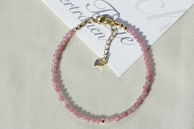 Op de afbeelding: Een delicate armband van roze edelstenen met een gouden sluiting en verlengketting. De armband is gemaakt van kleine, gefacetteerde kralen en heeft een hartvormige hanger.