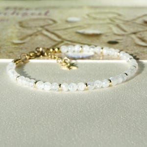 Pode incluir: Uma pulseira delicada com pequenas contas brancas translúcidas intercaladas com minúsculos espaçadores de metal dourado. A pulseira inclui um fecho lagosta dourado, uma corrente ajustável e um pequeno pingente em forma de estrela.