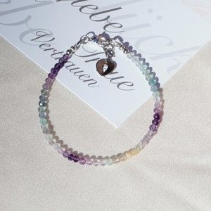 May include: A delicate bracelet made with small, colorful beads in shades of purple, blue, and green. The bracelet features a silver charm in the shape of a heart with a small cut-out.