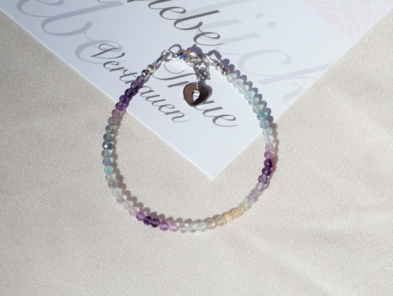 May include: A delicate bracelet made with small, colorful beads in shades of purple, blue, and green. The bracelet features a silver charm in the shape of a heart with a small cut-out.