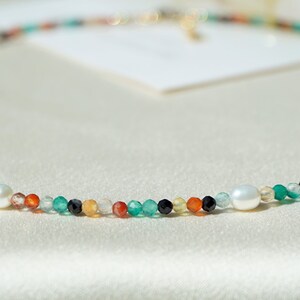 May include: A delicate necklace made with a variety of colorful beads, including green, orange, red, and black, interspersed with two white pearls.