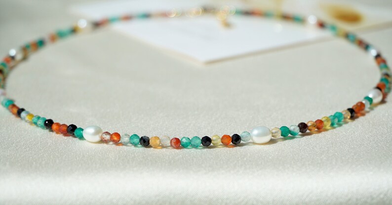 May include: A delicate necklace made with a variety of colorful beads, including green, orange, red, and black, interspersed with two white pearls.