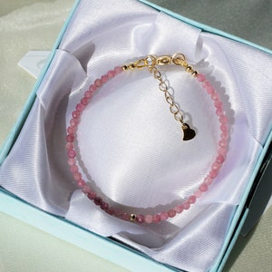 Op de afbeelding: Een delicate armband van roze edelstenen met een gouden hartbedel en een gouden sluiting. De armband wordt gepresenteerd in een lichtblauwe geschenkdoos met een witte satijnen voering.