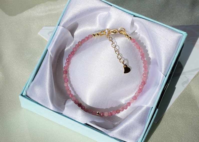 Op de afbeelding: Een delicate armband van roze edelstenen met een gouden hartbedel en een gouden sluiting. De armband wordt gepresenteerd in een lichtblauwe geschenkdoos met een witte satijnen voering.