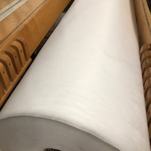 May include: A large roll of white fabric is wound around a wooden core. The fabric is smooth and appears to be non-woven.