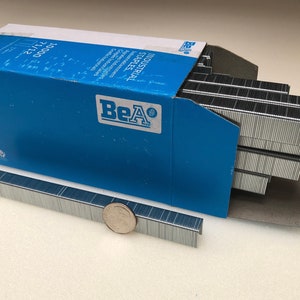 May include: A box of 10,000 BeA Industrial Staples, size 21/15, in a blue box with the BeA logo. The staples are silver and are arranged in rows in the box. A single row of staples is outside the box for size comparison.