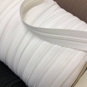 May include: A close-up of a spool of white zipper tape. The tape is wound tightly and the zipper teeth are visible.