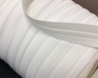 May include: A close-up of a spool of white zipper tape. The tape is wound tightly and the zipper teeth are visible.