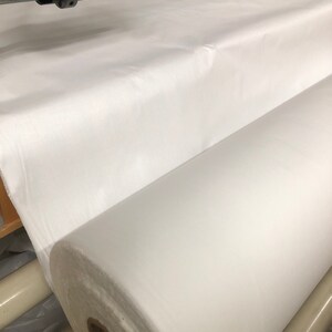 May include: A large roll of white fabric is being unrolled on a machine. The fabric is smooth and appears to be a type of cotton.