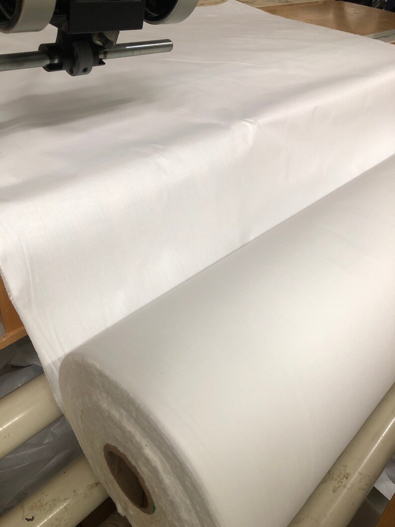 May include: A large roll of white fabric is being unrolled on a machine. The fabric is smooth and appears to be a type of cotton.