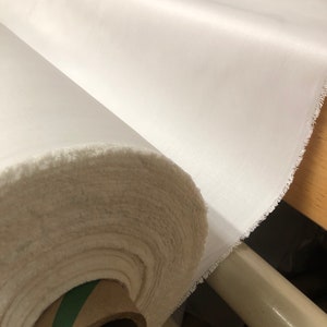 May include: A roll of white fabric with a green and brown cardboard core. The fabric is rolled up tightly and has a slightly frayed edge.