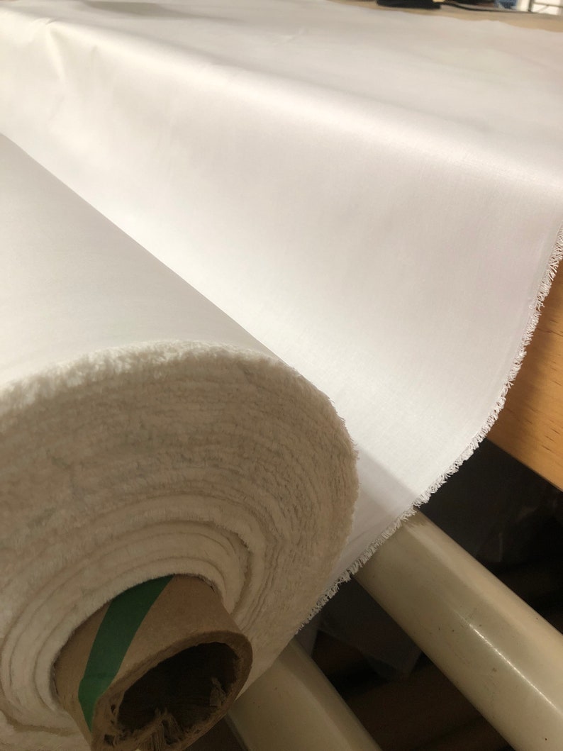 May include: A roll of white fabric with a green and brown cardboard core. The fabric is rolled up tightly and has a slightly frayed edge.
