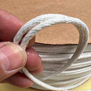 May include: A spool of white cotton cord, wound tightly and held in a hand. The cord is braided and has a smooth, matte finish.