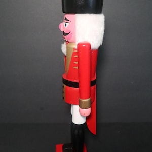 Peut inclure: Figurine de casse-noisette en bois au design classique. Il présente un manteau rouge avec des accents dorés, un chapeau noir et un col en fourrure blanche. La figurine repose sur une base rouge et a des bottes noires. Décoration de vacances traditionnelle.