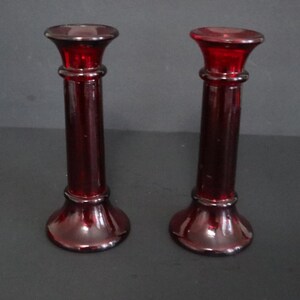 May include: A pair of ruby red glass candle holders. Each has a flared base, a cylindrical stem, and a flared top. The candle holders are made of transparent, glossy glass.