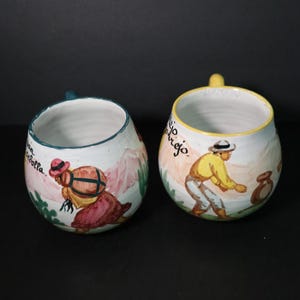 May include: Two ceramic mugs with hand-painted scenes. One mug depicts a person carrying a bundle, the other a person working in a field. The mugs have colourful designs and text, with blue and yellow handles.