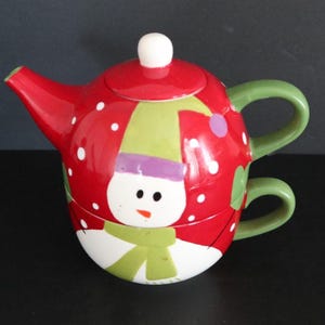 May include: A red and white snowman-themed teapot with a matching cup. The teapot features a snowman face with a green scarf and hat, and white polka dots. The handle and cup handle are green.