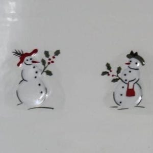 May include: White ceramic dish featuring six snowman illustrations. Each snowman is decorated with hats, scarves, and accessories in black, red, and green. The snowmen are drawn with black outlines and simple details.