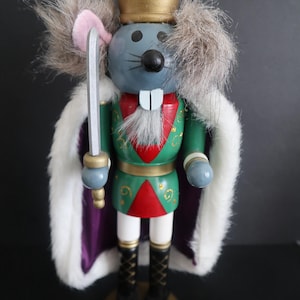 May include: A novelty nutcracker figurine of a mouse king. It wears a gold crown with red jewels, a green and red uniform, and a white fur cape. The mouse holds a silver sword and stands on a gold base.