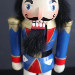 May include: A traditional wooden nutcracker figure. It has a red and gold crown, a blue and red uniform, a black moustache, and black hair. The figure stands on a red base.