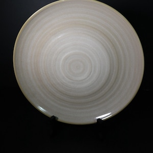 May include: A cream-coloured ceramic plate with a swirled pattern. The plate has a slightly raised rim with a light yellow border. The plate is round and appears handmade. The plate is on a black background.