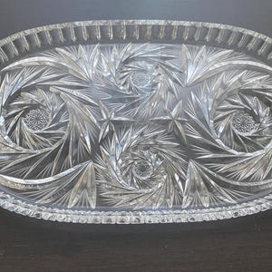 May include: Clear, oval crystal serving platter with detailed starburst and spiral designs. The tray has a textured rim and is suitable for serving food. A decorative item for special occasions. The dimensions are in centimetres.