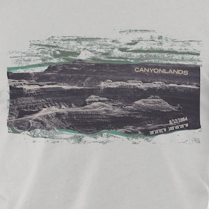 May include: White t-shirt featuring a graphic of Canyonlands National Park. The image shows a landscape with the text "CANYONLANDS" and the date "9/12/1964" along with coordinates. The design has a vintage, travel-inspired aesthetic.