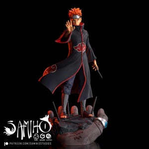 Peut inclure: Un personnage d'anime aux cheveux roux avec un symbole de nuage rouge sur sa robe noire se tient sur une base en forme de main. La base est gris foncé avec des accents rouges et le texte "Ce monde connaîtra la DOULEUR".