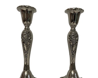 May include: Two silver-colored candlesticks with ornate detailing. The candlesticks have a flared base, a slender stem with a grape cluster design, and a ruffled top. The design is symmetrical, and the candlesticks are set against a white background.