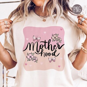 以下が含まれることがあります： クリーム色のTシャツ。筆記体で「Motherhood」の文字、蝶、花、ハートがピンク色の背景に描かれています。デザインはTシャツの中央に配置され、柔らかくフェミニンな美しさを醸し出しています。