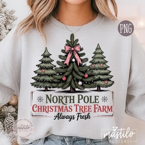 May include: A light gray sweatshirt featuring a Christmas tree design. The graphic includes three green Christmas trees, a pink bow, and ornaments. Text reads "North Pole Christmas Tree Farm Always Fresh."