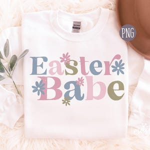 Może przedstawiać: Biała bluza z nadrukiem "Easter Babe" w różowych, niebieskich i zielonych literach z kwiatowymi akcentami.