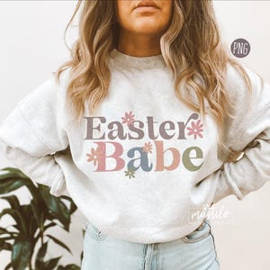 Może przedstawiać: Biała bluza z napisem "Easter Babe" w kolorowych, retro literach i kwiatowymi akcentami.