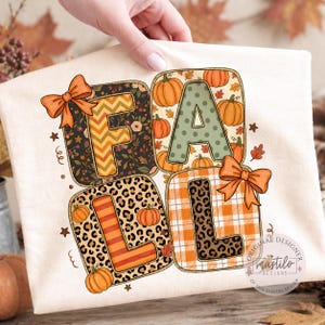 May include: Cream-coloured t-shirt featuring the word "FALL" in a square design. Each letter is filled with a different pattern, including floral, leopard print, and plaid. Orange bows and pumpkins decorate the design, perfect for autumn.