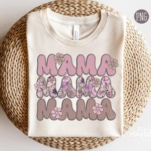 May include: A beige t-shirt with the word "Mama" repeated three times in pink, brown, and pink lettering. Each word is decorated with floral designs in shades of pink and brown.