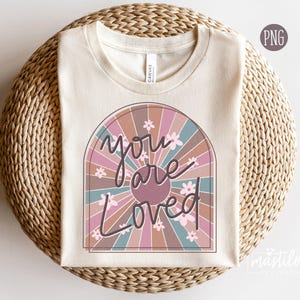 May include: A beige t-shirt featuring a retro-style sunburst design in shades of pink, brown, and blue. The design includes the text "You are Loved" in a stylised font.