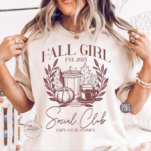 以下が含まれることがあります： FALL GIRL EST. 2025 Social Club COZY CUTE COMFYという文字と、カボチャ、カップ、マグカップ、秋の葉が2本の枝に囲まれたデザインが描かれた、ダークブラウンのグラフィック入りベージュTシャツ。