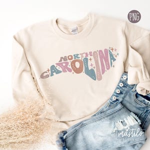 May include: A cream-colored sweatshirt with a graphic design of the state of North Carolina in pink, blue, and brown with the words "NORTH CAROLINA" in a retro font. The sweatshirt is laying on a light brown surface with a pair of blue jeans.