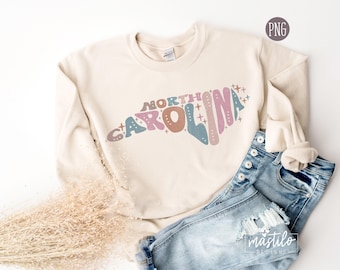 May include: A cream-coloured sweatshirt with a graphic design of the state of North Carolina in pink, blue, and brown with the words "NORTH CAROLINA" in a retro font. The sweatshirt is lying on a light brown surface with a pair of blue jeans.