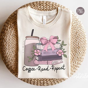 以下が含まれることがあります： コーヒーカップ、本、ピンクのリボンが描かれたベージュのTシャツ。グラフィックの下に「Coffee. Read. Repeat.」の文字が印刷されています。