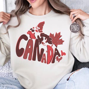 May include: A cream-colored sweatshirt with a red and brown graphic of the word "CANADA" with a stylized outline of the country's map and a red maple leaf.