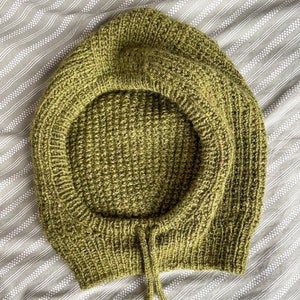 May include: A knitted hood in a muted olive green color. The hood features a drawstring closure and a ribbed knit design around the face opening. The hood is made of a textured yarn and is laying on a striped fabric.