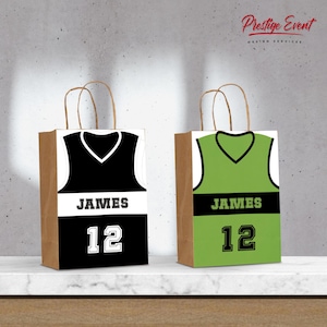 Peut inclure: Deux sacs cadeaux en papier kraft avec des poignées noires. Chaque sac est imprimé avec un motif de maillot de basket-ball noir et blanc et un maillot de basket-ball vert et noir. Les maillots portent le nom "JAMES" et le numéro "12" imprimé dessus.