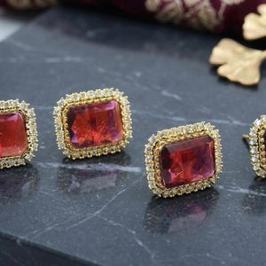 May include: Four gold-toned earrings with rectangular, deep red gemstones. Each gem is surrounded by a halo of small, clear stones. The earrings are displayed on a dark, textured surface.