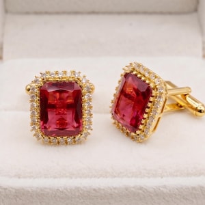 Puede incluir: Un par de gemelos dorados con una gran piedra roja rectangular facetada en el centro. Cada piedra está rodeada por un halo de pequeñas piedras claras y brillantes engastadas en un marco de metal de color dorado con cierre de palanca.