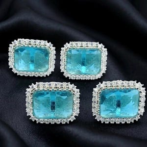 Puede incluir: Cuatro pendientes cuadrados con una piedra central azul claro y un borde incrustado de color plateado. Los pendientes se exhiben sobre una tela negra y sedosa, creando un contraste que resalta los detalles.